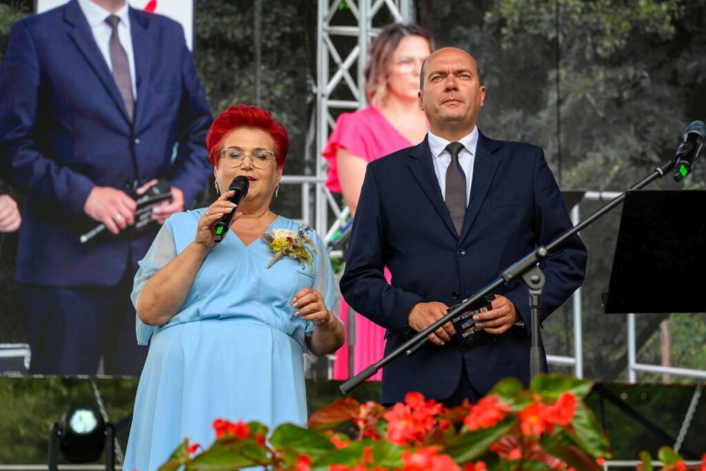 Dwie osoby w eleganckim stroju występują na scenie. Kobieta w błękitnej sukni trzyma mikrofon. Tło to ekran z zielenią i kwiatami.