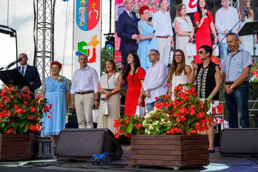 Grupa ludzi stoi na scenie, tło stanowi duży ekran z ich wizerunkami. Scena ozdobiona kolorowymi kwiatami. Widać jasny dzień.