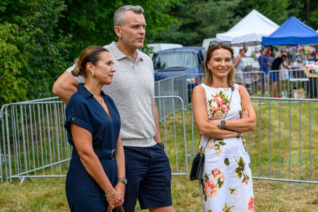 Trzy osoby stoją na trawie. Dwie kobiety, ubrane w kwiecistą sukienkę i granatowy strój, oraz mężczyzna w jasnej koszulce. W tle namioty i ogrodzenie.