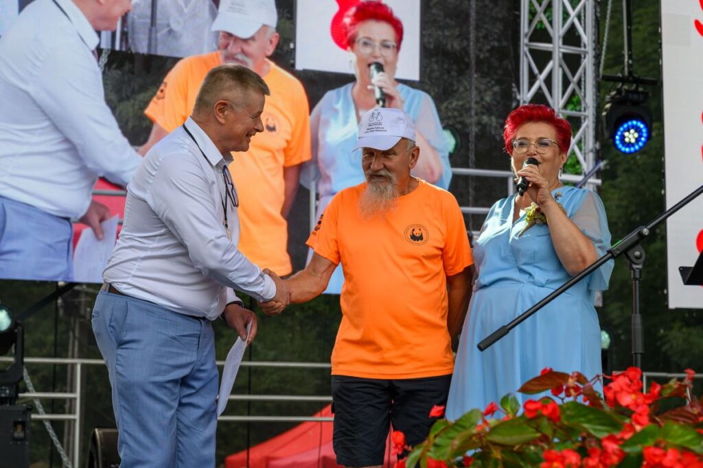 Na zdjęciu trójka ludzi na scenie: mężczyzna w białej koszuli, mężczyzna w pomarańczowej koszulce i kobieta w niebieskiej sukni. Mężczyźni podają sobie ręce, kobieta mówi do mikrofonu. Tło stanowi ekran z ich pow