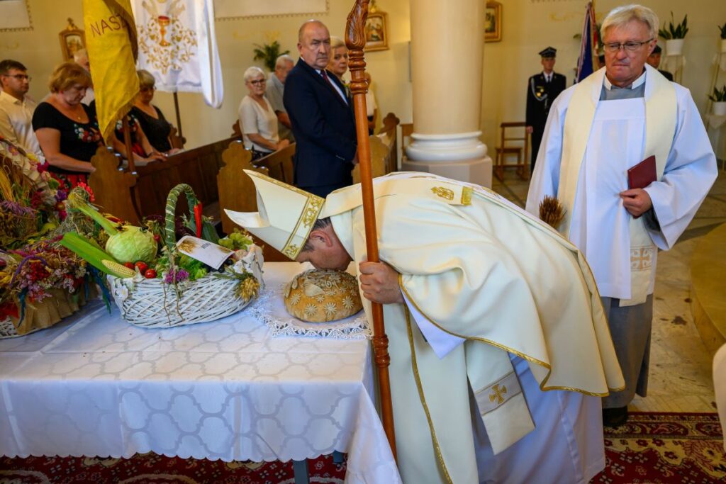 Na zdjęciu widać uroczystość religijną w kościele. Kapłan w białej szacie z pastorałem pochylony nad chlebem na ozdobnym stole. W tle wierni i dekoracje z koszami owoców.