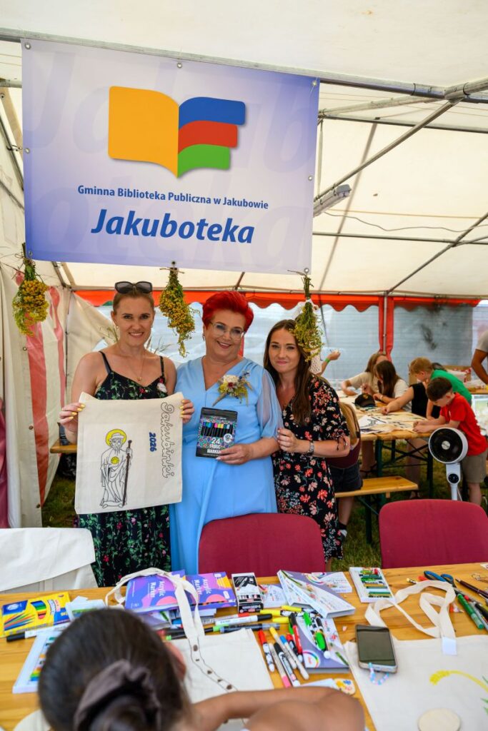Zdjęcie przedstawia trzy kobiety w namiocie biblioteki. Jedna z nich trzyma torbę z rysunkiem, druga książkę. W tle są dzieci przy stołach.