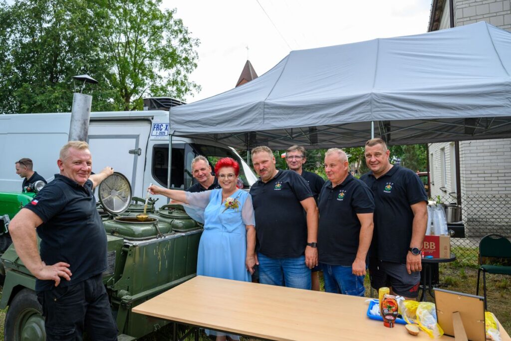 Grupa ludzi stoi przy polowej kuchni wojskowej na zielonym tle. Kobieta w niebieskiej sukience miesza potrawę. Namiot i samochód w tle.
