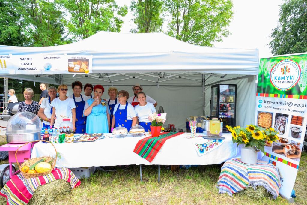 Grupa osób w niebieskich fartuchach stoi przed namiotem, na stole ciasta i napoje. W tle zielone drzewa. Obok plakat z logiem.