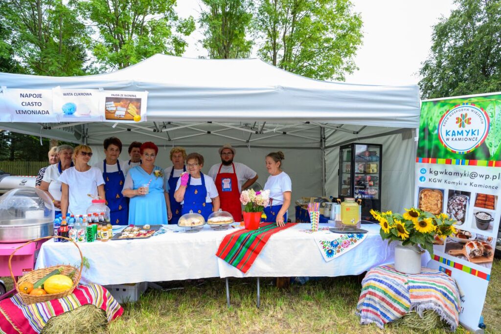 Stoisko na festynie z grupą osób w niebieskich fartuchach. Na stole napoje, ciasta i dekoracje kwiatowe. Tło stanowi zielone otoczenie i biały namiot.