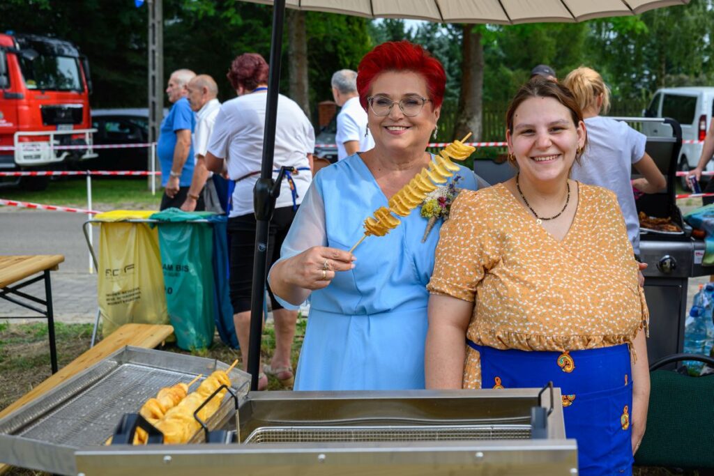 Dwie uśmiechnięte kobiety stoją przy frytkownicy na festynie. Jedna trzyma spiralne frytki na patyku. W tle ludzie i czerwony wóz strażacki.