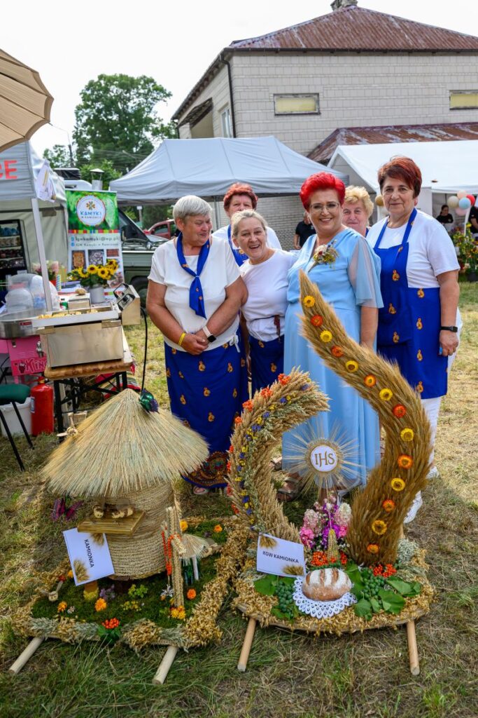 Grupa kobiet w niebiesko-białych strojach stoi na tle straganów. Przed nimi ozdoby z kłosów i słomy, kolorowe kwiaty. Tło to wiejskie targi z białymi namiotami.
