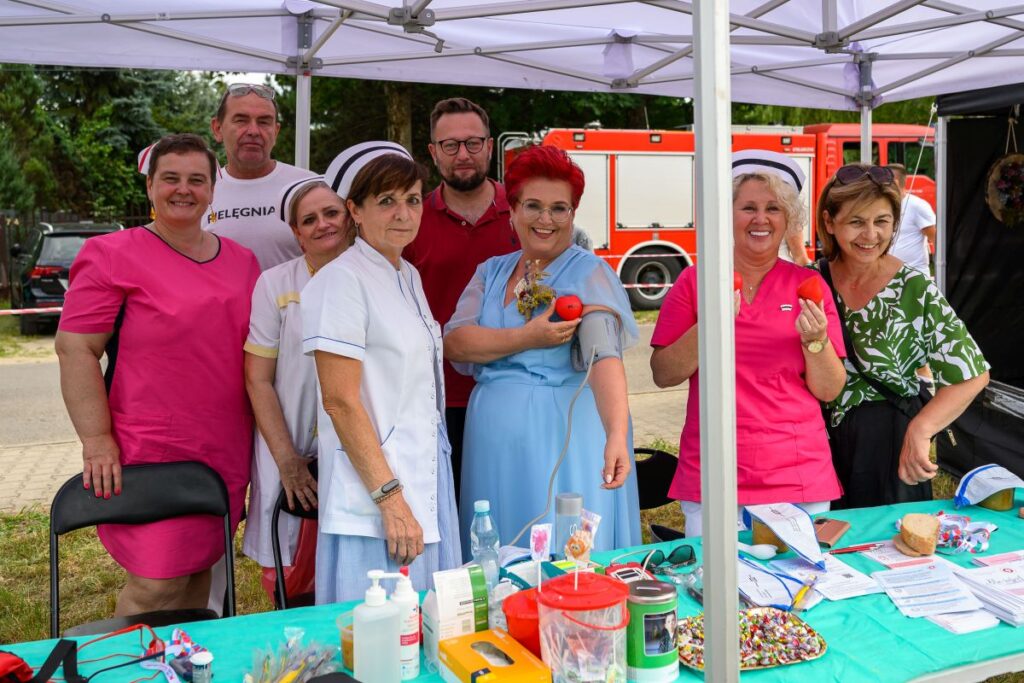 Grupa osób w białych i różowych ubraniach medycznych stoi pod namiotem. Tło to zielone drzewa i czerwony pojazd. Na stole leżą artykuły medyczne i dokumenty.