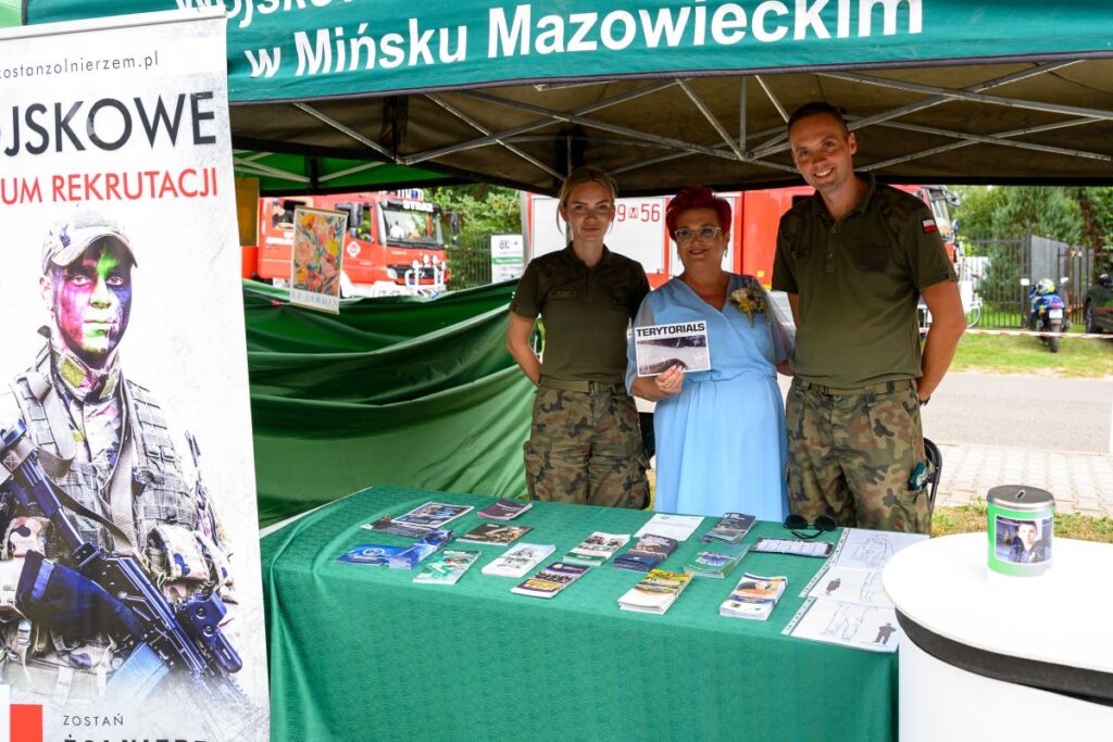 Na zdjęciu jest zielony namiot z napisem, stół z ulotkami i trzy osoby w mundurach. W tle czerwone pojazdy strażackie. Dominuje kolor zielony i czerwony.
