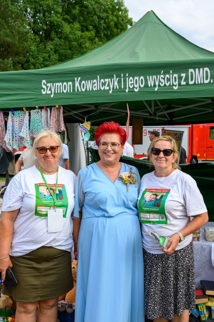 Na zdjęciu są trzy uśmiechnięte osoby pod zielonym namiotem z napisem. Dwie z nich noszą białe koszulki z grafiką. W tle widać elementy stoiska i drzewa.