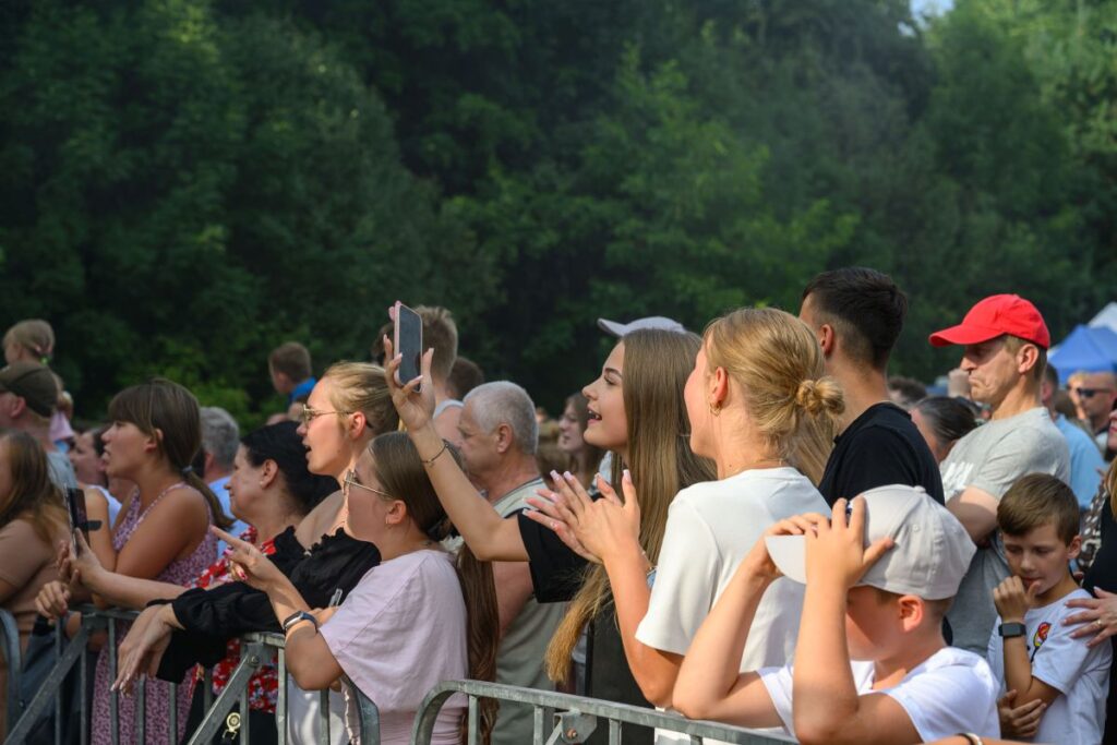 Grupa ludzi na koncercie, uśmiechnięte twarze, zielone drzewa w tle, różnorodność kolorowych ubrań i emocjonująca atmosfera.