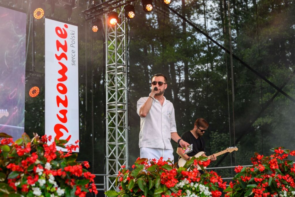 Mężczyzna w okularach na scenie śpiewa z mikrofonem, obok niego gitarzysta. W tle drzewa, przed sceną czerwone kwiaty. Na lewo baner z napisem Mazowsze.