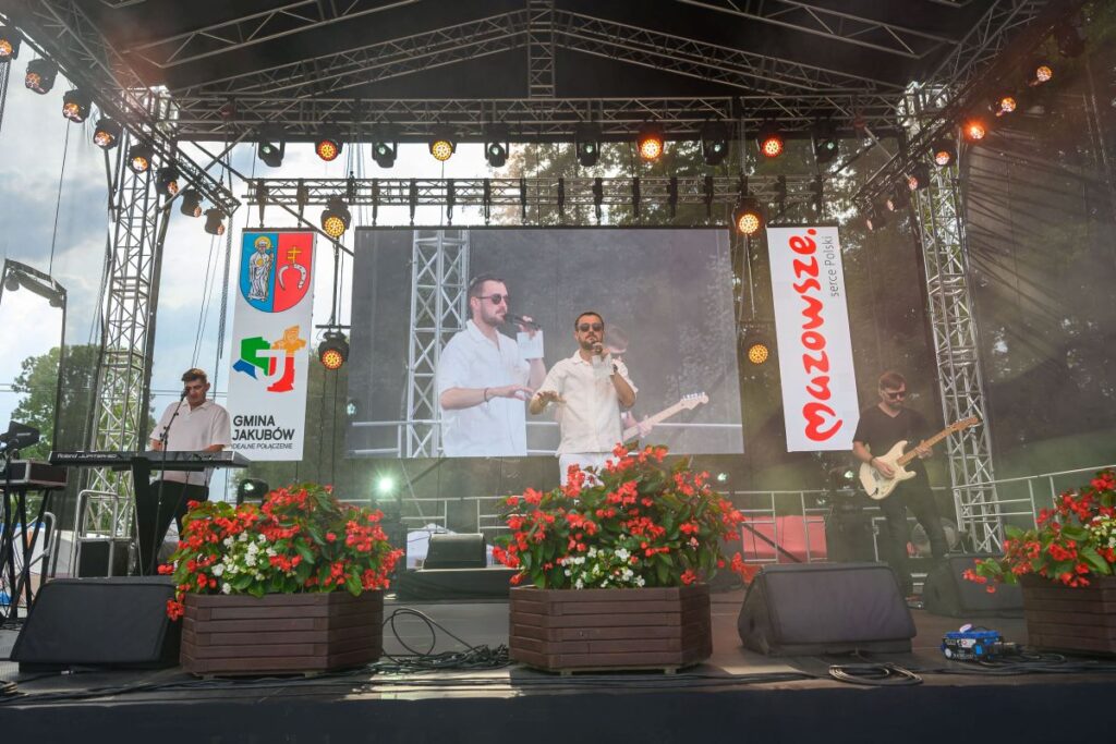 Na scenie zespół muzyczny występuje pod zadaszeniem. Z przodu czerwone i białe kwiaty, w tle reklamy Mazowsza. Centralnie widoczny ekran z występem.