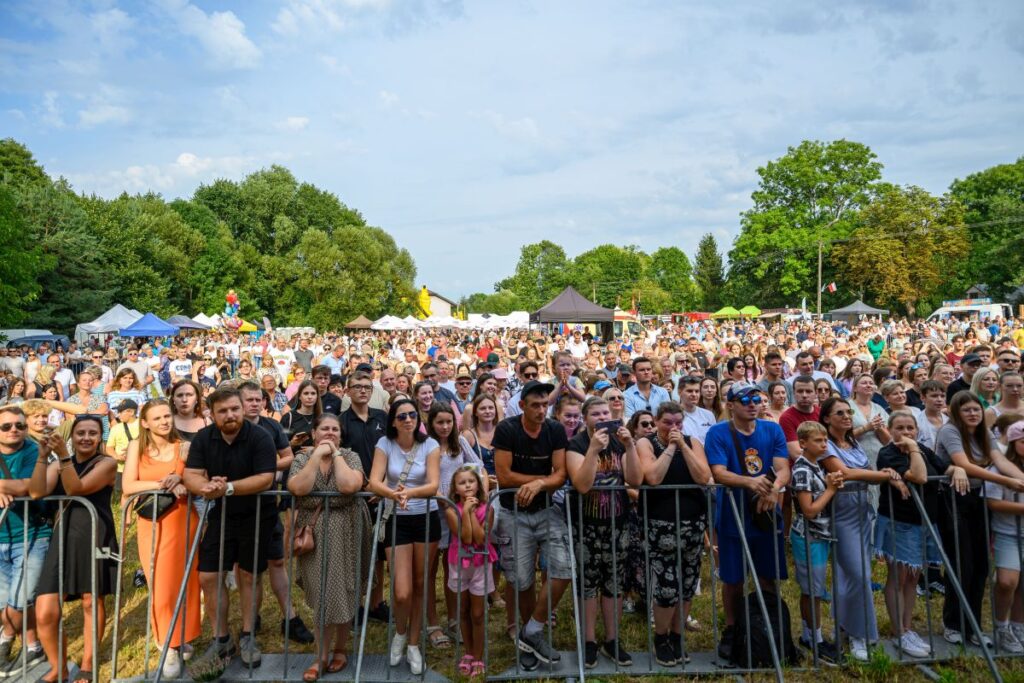 Tłum zgromadzony na otwartych przestrzeniach festiwalu, otoczony przez zielone drzewa. Ludzie są radośni i patrzą w stronę sceny. W tle widać namioty i stoiska.
