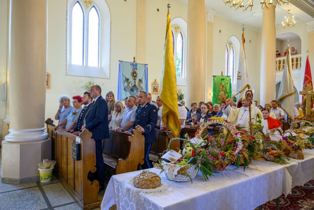 Wnętrze kościoła z witrażami, ludzie stoją przy drewnianych ławkach. Na stole białe obrusy i dekoracje z dożynkami. Jasne ściany, wysoki sufit i kolumny.