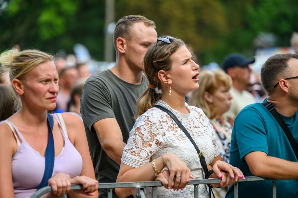 Grupa ludzi stoi za barierkami na zewnątrz. Tło jest rozmyte, zielone drzewa. Kobiety i mężczyźni wyglądają na skupionych.
