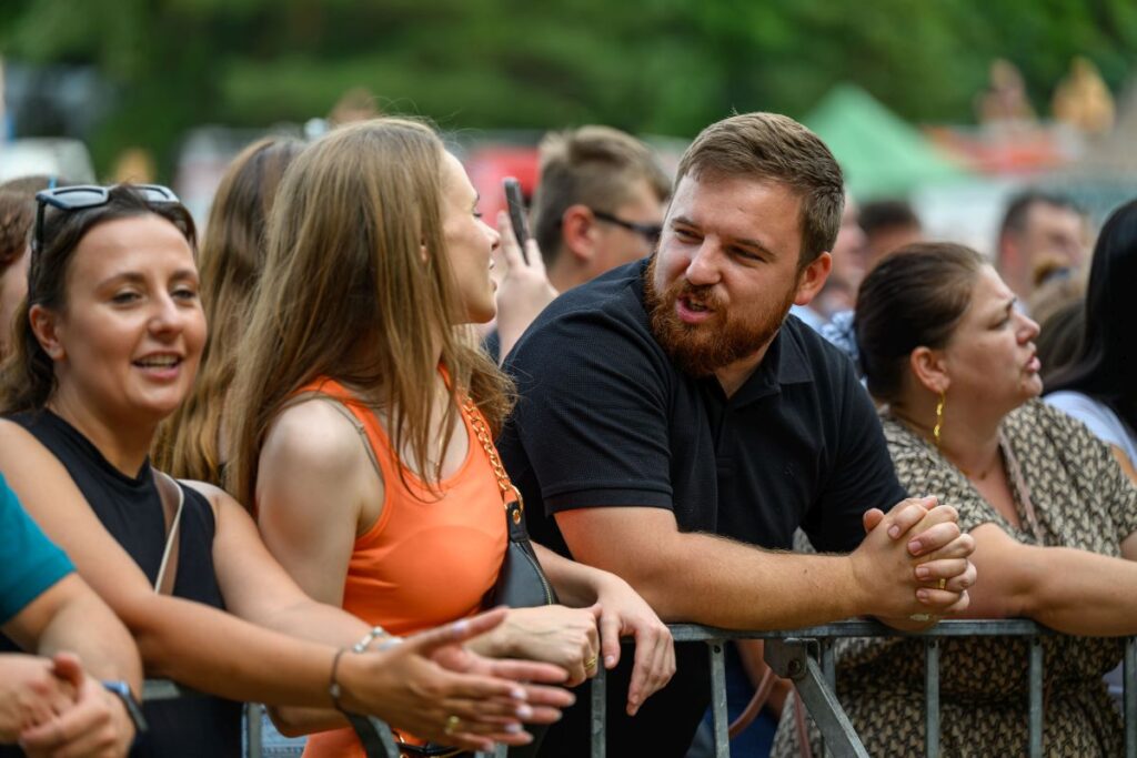 Grupa ludzi stoi przy barierce na zewnątrz, rozmawiając i uśmiechając się. Kobieta w pomarańczowej bluzce i mężczyzna w czarnej koszulce tłumnie zgromadzeni. W tle rozmyte, zielone drzewa i nami
