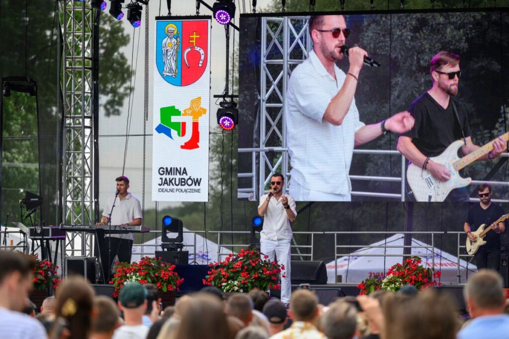 Mężczyzna śpiewa na scenie, obok gitarzysta. Tło to duży ekran z transmisją występu. Na lewo baner Gmina Jakubów.