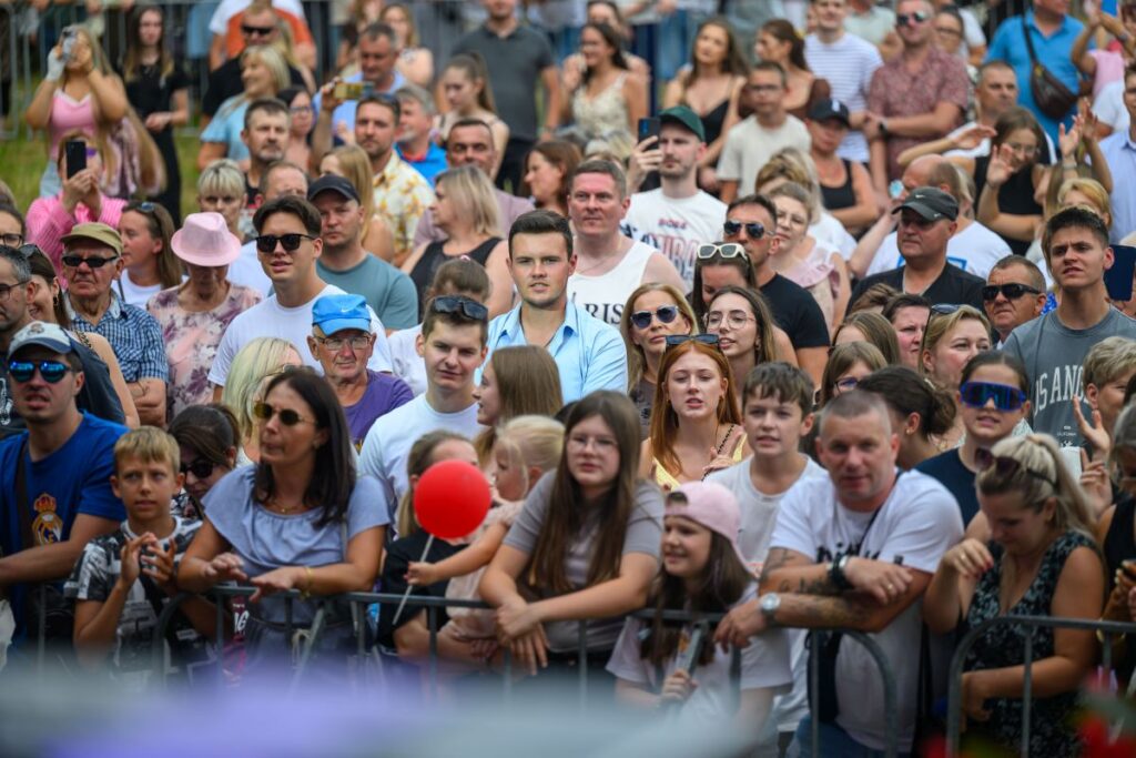 Tłum w różnorodnym ubraniu gromadzi się, patrząc w stronę sceny. Ludzie w okularach przeciwsłonecznych, jasne i ciemne kolory. W tle płot, niebo niewidoczne.