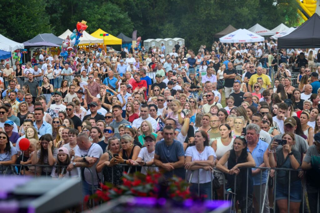 Na zdjęciu widać tłum ludzi na festiwalu na świeżym powietrzu. Tła stanowią kolorowe namioty i zielone drzewa. Atmosfera jest żywa i pełna energii.