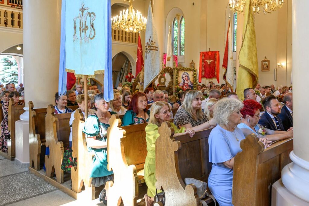 Wnętrze kościoła z wiernymi siedzącymi w drewnianych ławkach. Kolorowe sztandary i obrazy religijne ozdabiają tło. Jasne światło wpada przez okna, oświetlając scenę.