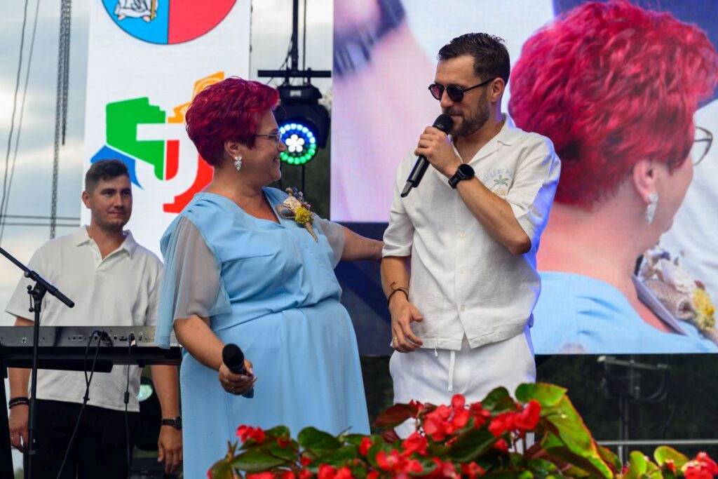 Na scenie kobieta w niebieskiej sukience i mężczyzna w białej koszuli rozmawiają. W tle duży ekran oraz logo. Na pierwszym planie czerwone kwiaty.
