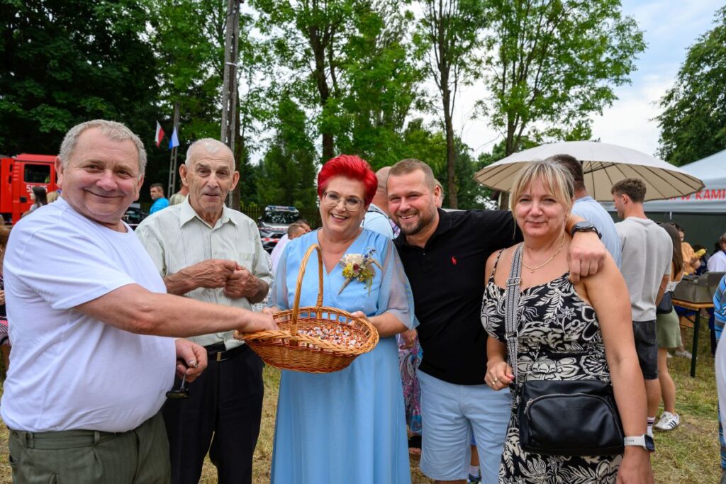 Grupa ludzi uśmiecha się, trzymając koszyk na tle drzew. Kobieta w niebieskiej sukni stoi pośrodku. Dzień jest słoneczny.