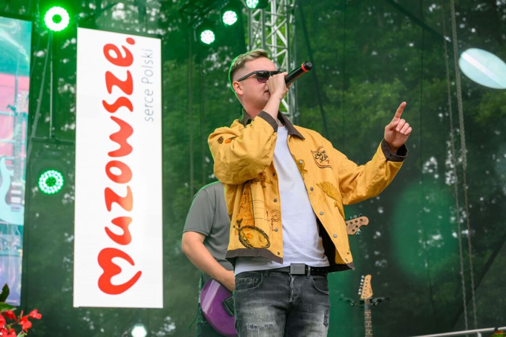 Mężczyzna w żółtej kurtce śpiewa na scenie, trzymając mikrofon. W tle zielone drzewa i duży biały transparent z czerwonym napisem. Obok niego widać gitarzystę.