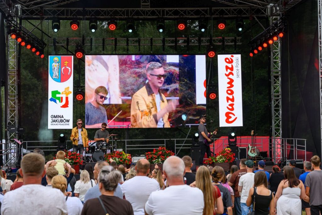 Koncert na scenie plenerowej, zespół gra przy tłumie ludzi. Na ekranie duże zbliżenie wokalisty w złotej kurtce. Kolorowe światła i kwiaty na scenie.