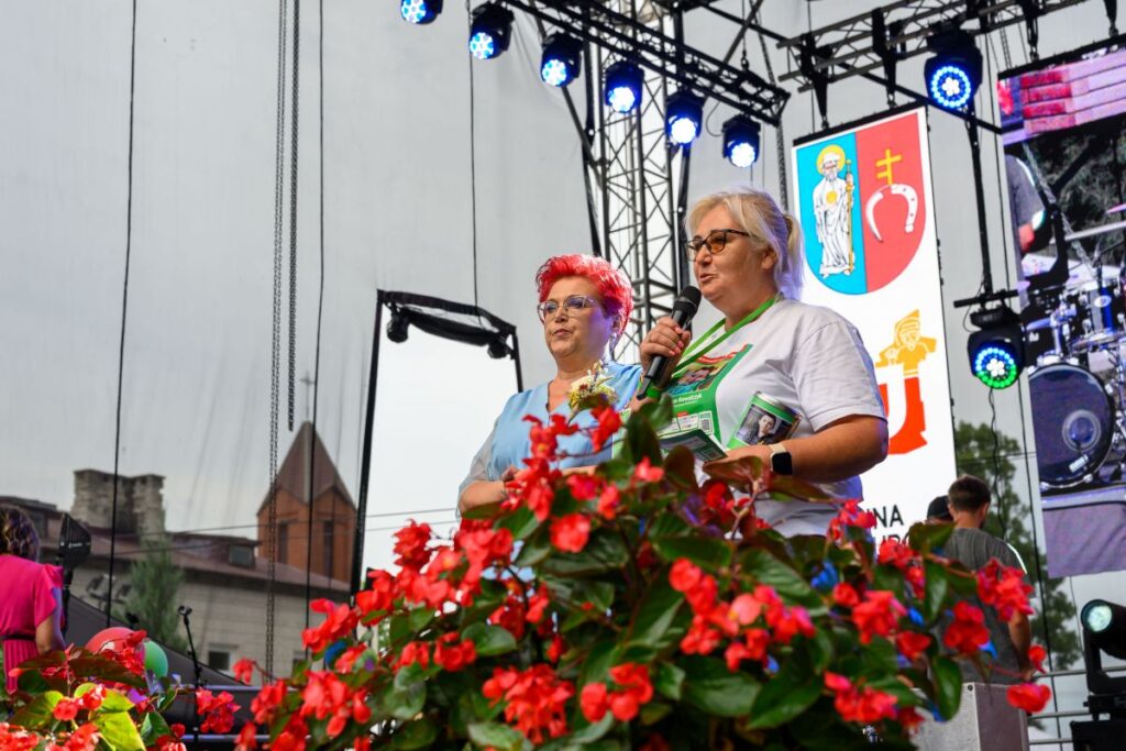 Na scenie dwie osoby przemawiają do mikrofonu. Przed nimi czerwone kwiaty, w tle herby i sprzęt muzyczny. Sceneria koncertowa z oświetleniem.