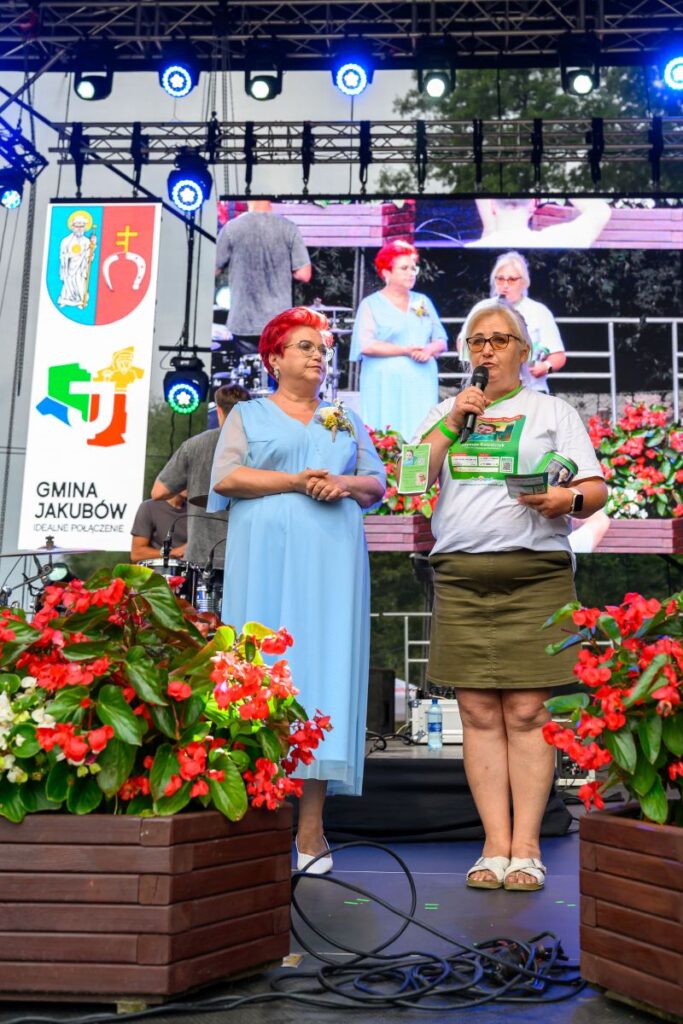 Na scenie stoją dwie kobiety, jedna w niebieskiej sukni, druga w białej koszulce, trzymają mikrofon. Tło to ekran z powiększonym ich obrazem, otoczonej czerwonymi kwiatami. Z boku widoczny jest baner z herbem