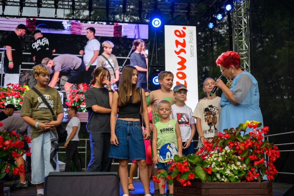 Na scenie znajduje się grupa dzieci i kobieta w niebieskiej sukni, trzyma mikrofon. W tle ekran z ludźmi i kwiatami. Obok baner z napisem.