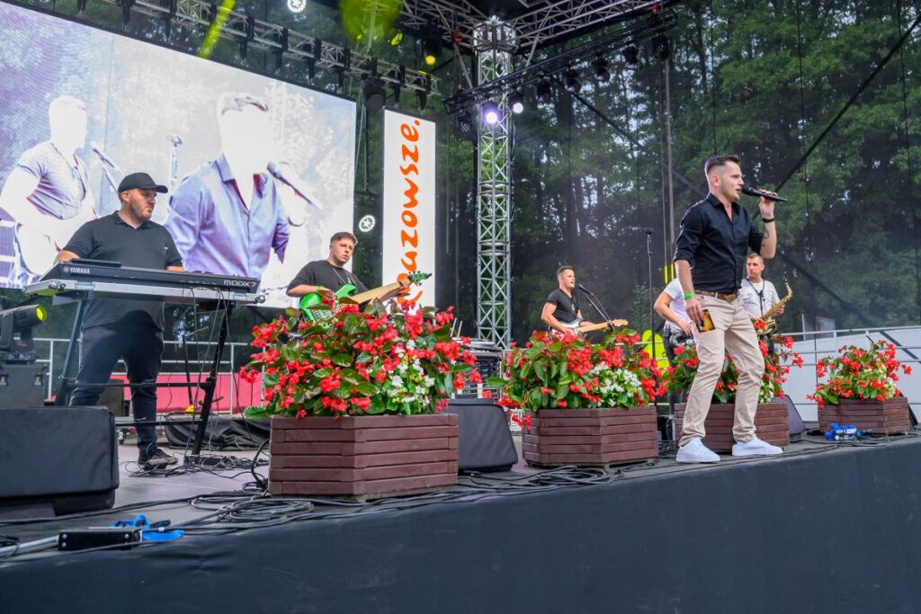 Na zdjęciu widać muzyków grających na scenie z instrumentami, otoczonych czerwonymi kwiatami. Duży ekran pokazuje ich w tle. Dookoła widoczny jest zalesiony krajobraz.