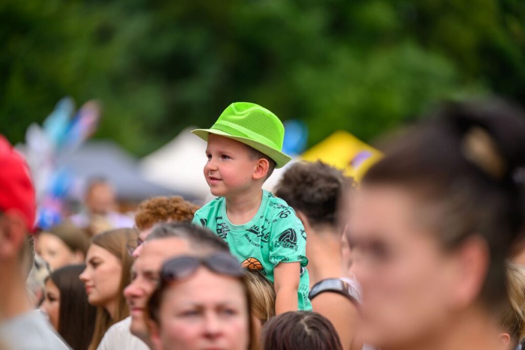 Chłopiec w zielonym kapeluszu siedzi na ramionach w tłumie. Ma na sobie zieloną koszulkę z nadrukiem. W tle rozmyta zieleń i namioty.