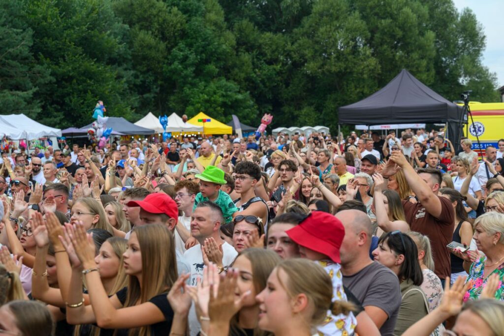Na zdjęciu tłum ludzi na świeżym powietrzu, wielu z podniesionymi rękami. W tle namioty i zielone drzewa. Kolorowe akcenty, głównie czerwone i zielone.