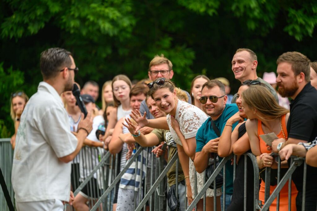 Tłum przed barierkami słucha mężczyzny w białej koszuli z mikrofonem. Osoby w odzieży letniej, uśmiechnięte. Tło tworzą zielone drzewa.