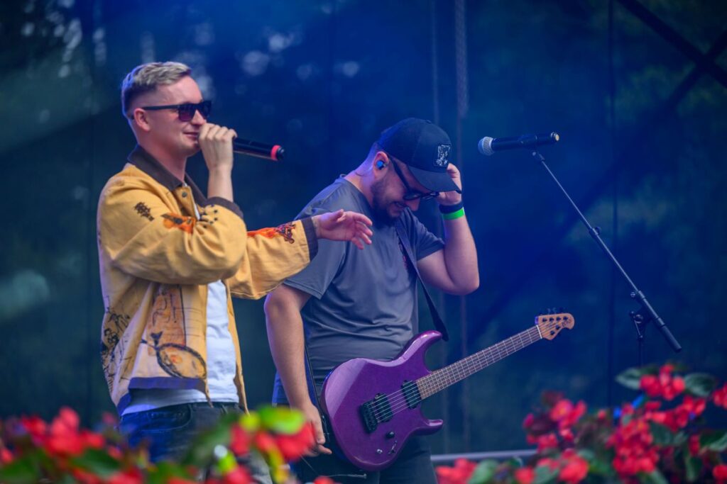 Na scenie dwaj mężczyźni: jeden z mikrofonem, drugi z gitarą. Pierwszy w jasnej kurtce, drugi w czapce. Tło: ciemne, kwiaty z przodu.