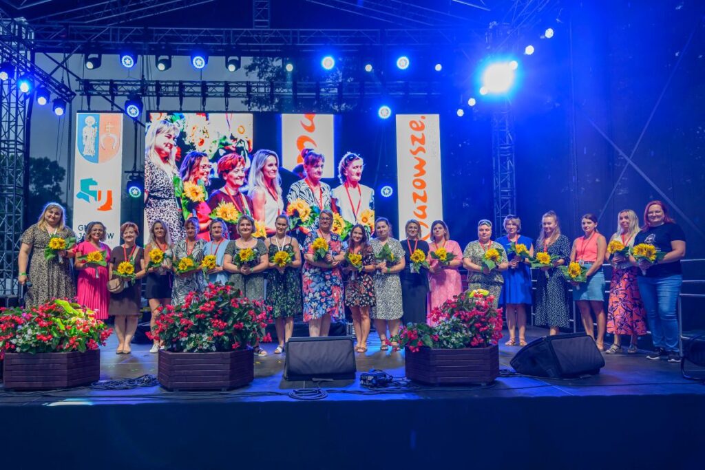 Na scenie stoi grupa kobiet z bukietami słoneczników. Tło jest ciemne, a za nimi widać jasne ekrany z napisami. Na scenie ozdobne kwiaty w donicach.