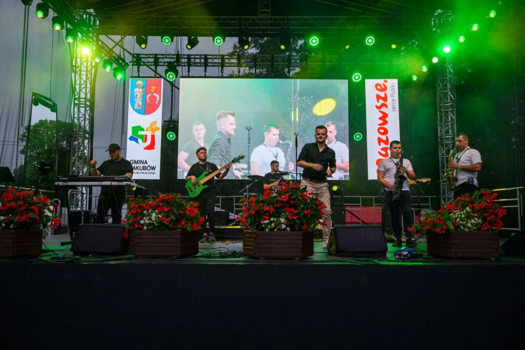 Grupa muzyczna występuje na scenie z zielonym oświetleniem. W tle widoczny jest duży ekran z wideo. Przed sceną znajdują się kwiaty w donicach.