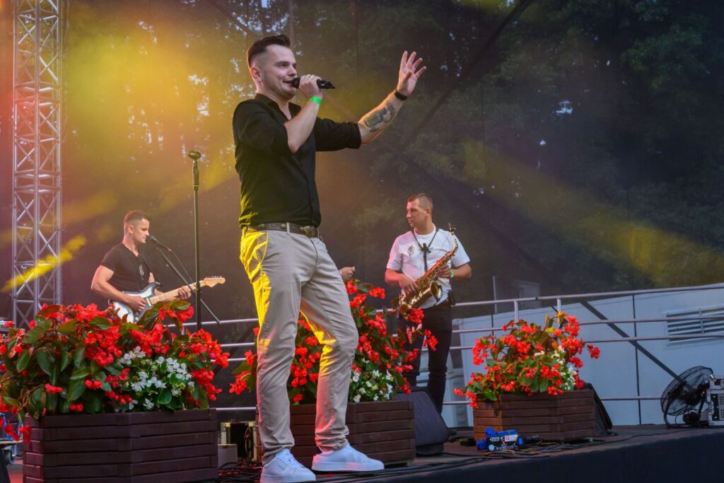 Na scenie stoją trzej mężczyźni z instrumentami muzycznymi. Jeden z nich śpiewa do mikrofonu, otoczony czerwonymi kwiatami. Tło jest ciemne, oświetlone żółtymi reflektorami.