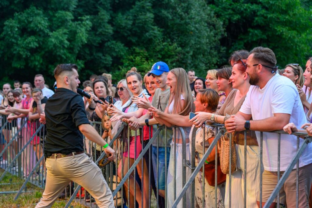 Na zdjęciu widać piosenkarza w czarnej koszuli i jasnych spodniach, śpiewającego dla tłumu. Publiczność, za barierką, reaguje entuzjastycznie. Tło to zielone drzewa, sugerujące plenerowy koncert.