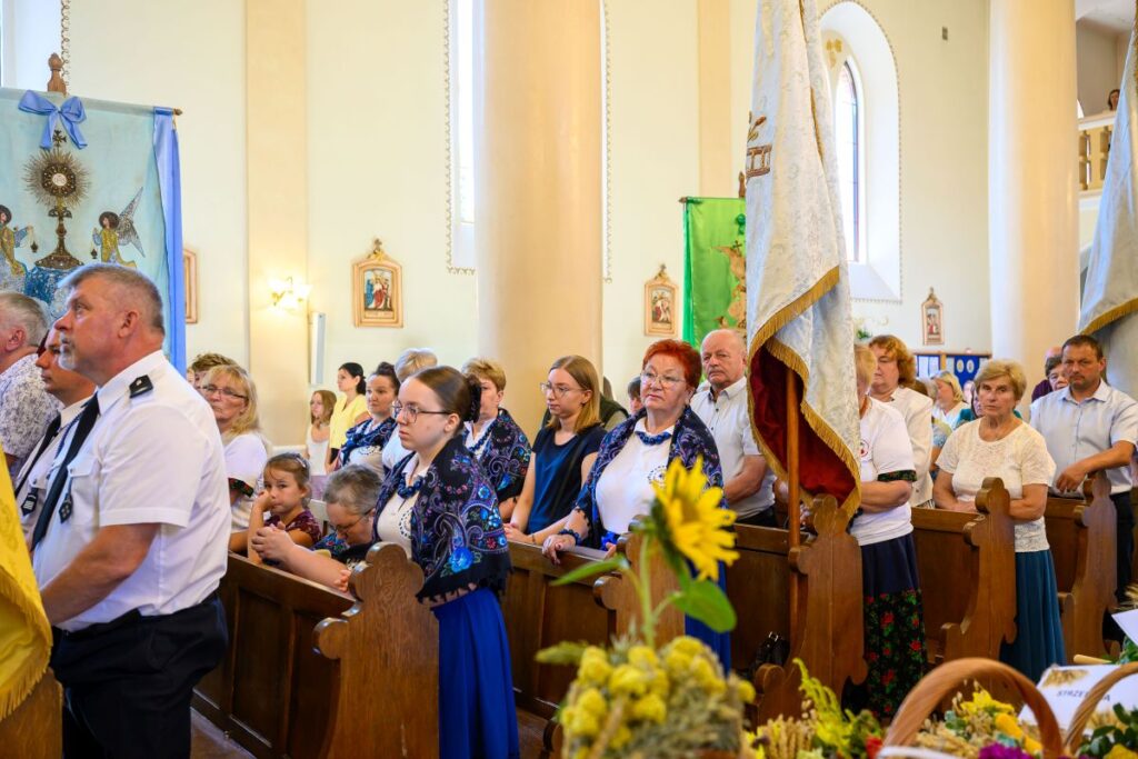 Grupa osób w kościele uczestniczy we mszy. Ubrani są w tradycyjne, kolorowe stroje, widoczne są dekoracyjne sztandary. W tle widać jasne ściany i ławki, z przodu ozdobne kwiaty.