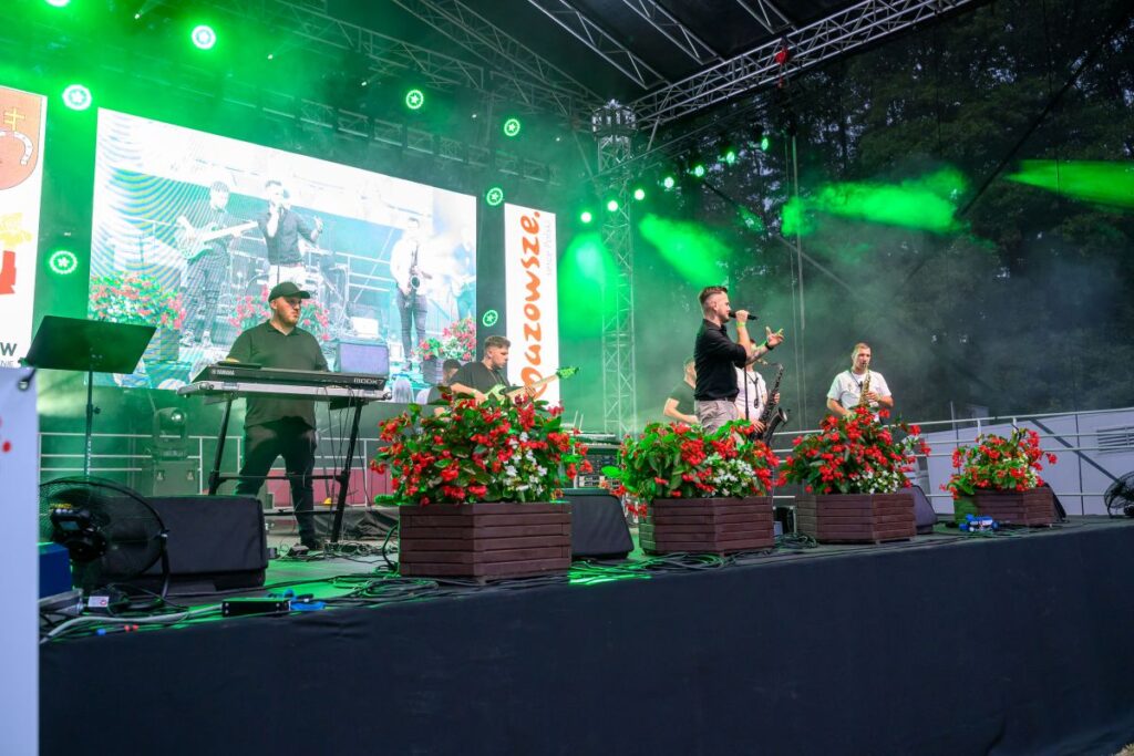 Grupa muzyczna występuje na scenie pełnej zielonych świateł i dymu. W tle duży ekran pokazujący muzyków. Przed sceną czerwone kwiaty w drewnianych donicach.