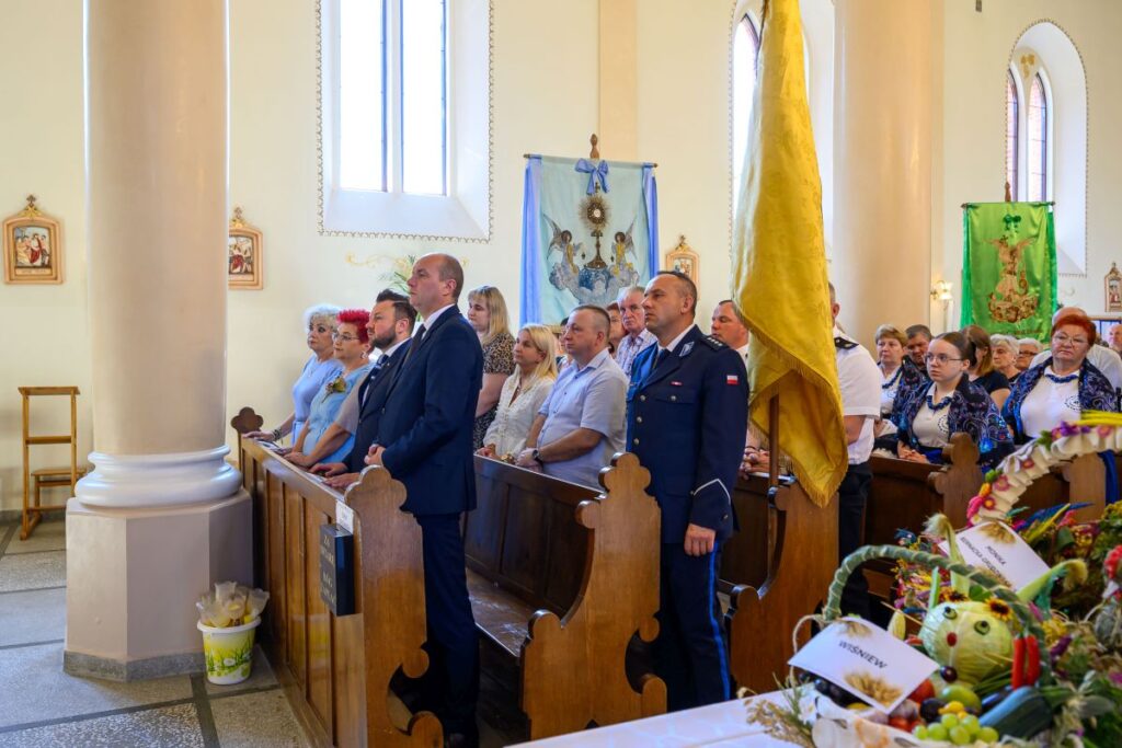 Grupa osób w kościele w eleganckich strojach. Ściany jasne, ozdobione kolorowymi sztandarami i ikonami. Na pierwszym planie kosze z dekoracjami.