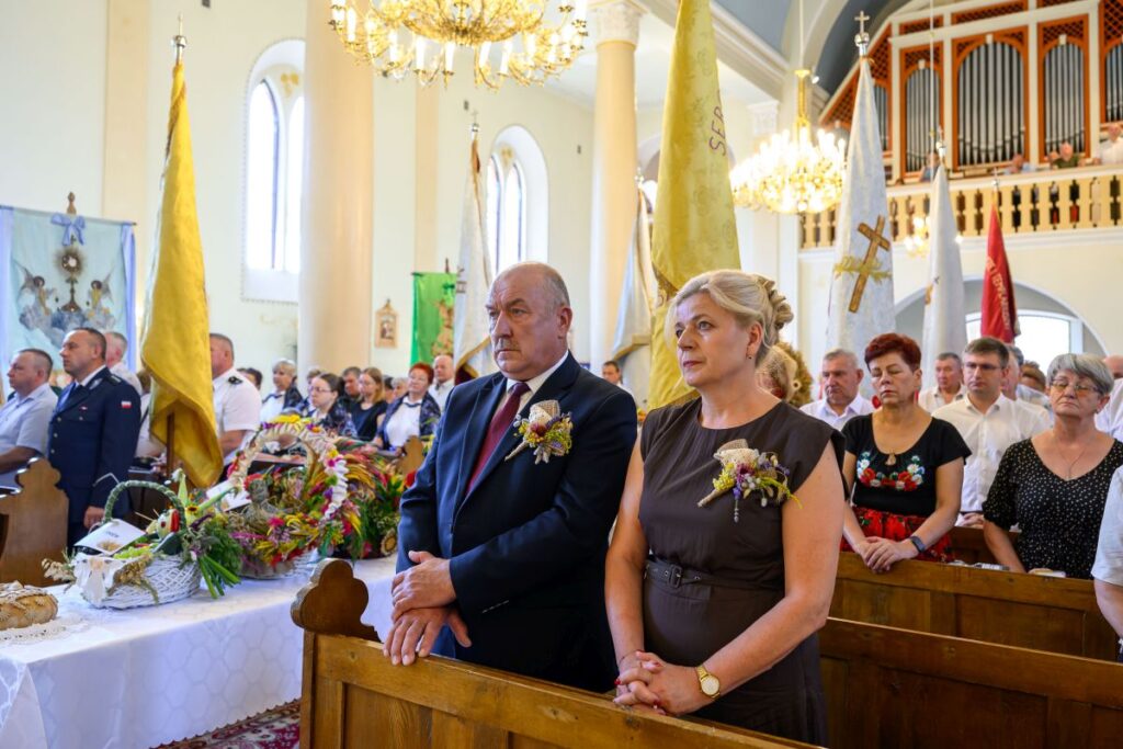 W jasnym kościele stoją ludzie z wieńcami, w tym para ubrana elegancko, w tle są flagi i organowy balkon.