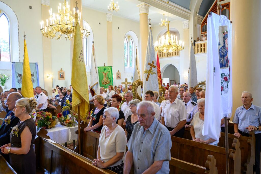 Grupa ludzi w kościele podczas ceremonii z flagami. Wnętrze jasne, z dużymi oknami i złotymi żyrandolami. Uczestnicy w białych i ciemnych strojach.