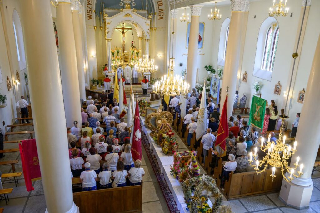 Wnętrze kościoła z wiernymi podczas mszy. Kolumny i żyrandole w kremowych odcieniach. Ołtarz zdobiony, na ścianach obrazy.