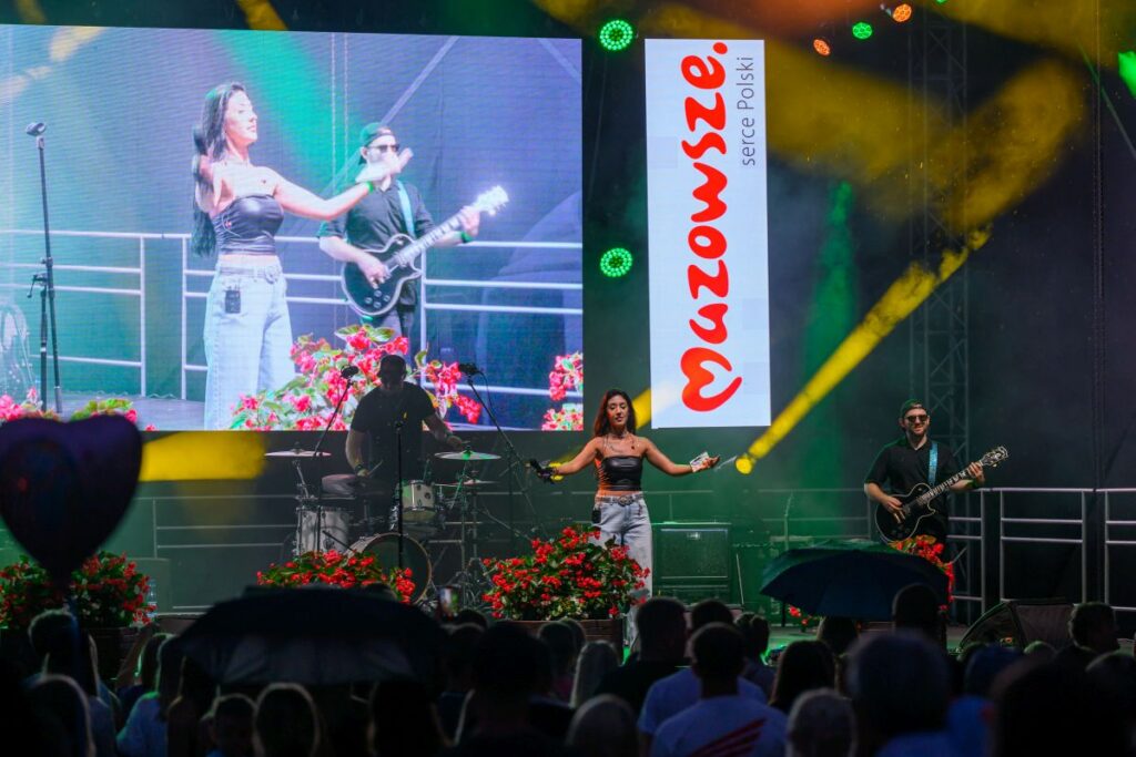 Na scenie występuje zespół muzyczny przed dużym ekranem z kolorowym logo Mazowsza. W tle widać zielone i żółte światła sceniczne. Publiczność ogląda koncert pod otwartym niebem.