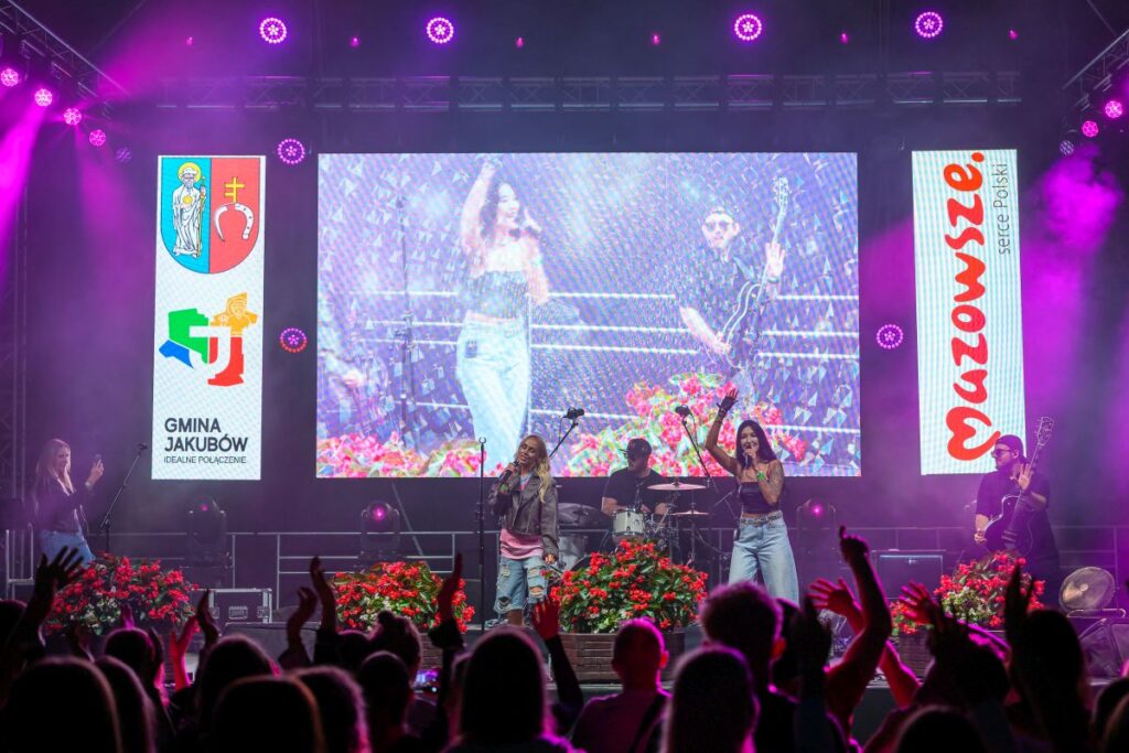 Koncert na scenie z zespołem i widownią. Jasne ekrany LED, fioletowe światła, kwiaty czerwone. Tła z logami Mazowsza i Jakubowa.