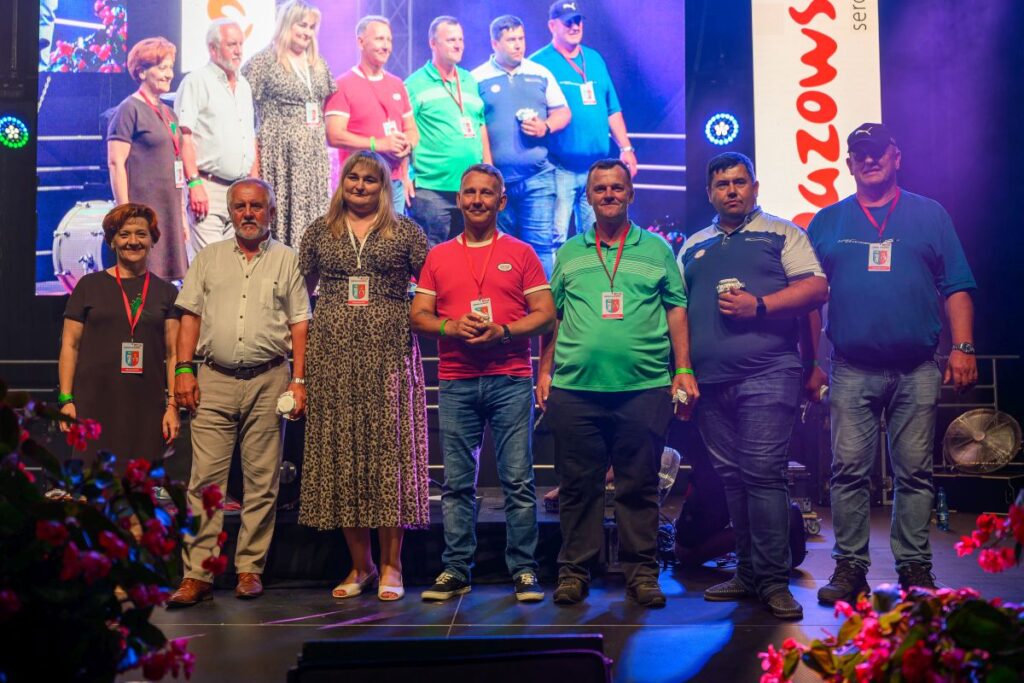 Grupa ludzi pozuje na scenie w kolorowych strojach. W tle jasne reflektory i duży ekran. Po bokach czerwone kwiaty.